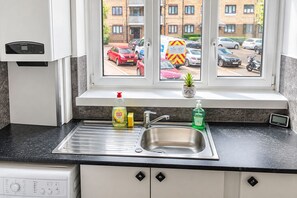 A kitchen with a stainless steel sink, a dish soap bottle, a hand soap bottle, and a small potted plant on the windowsill. - Bright 3 Bed flat with Balcony near Finsbury Park (Harringay)