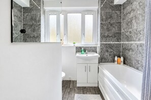 Ein modernes Badezimmer mit einer Badewanne, einem Waschbecken mit Pflegeprodukten und einem Fenster darüber.