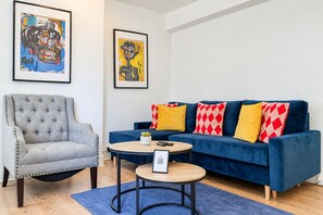 Ein Wohnzimmer mit einem grauen, gesteppten Sessel, einem blauen Sofa mit roten und gelben Kissen und einem runden Couchtisch. An der Wand hängen zwei gerahmte Kunstwerke.