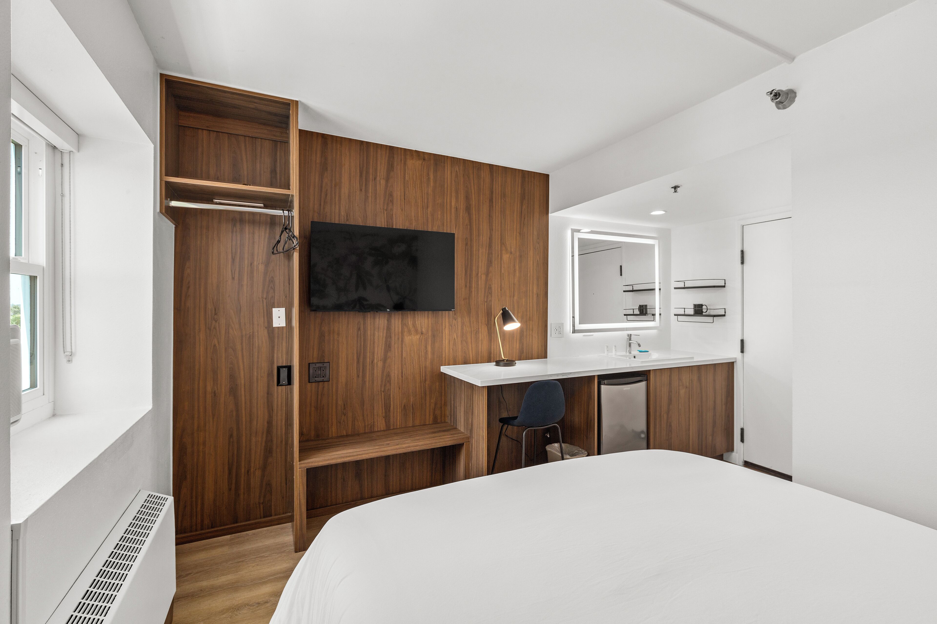 A modern hotel room with a wooden wardrobe, a bed, a desk with a chair, a TV, and a bathroom with a mirror and sink.