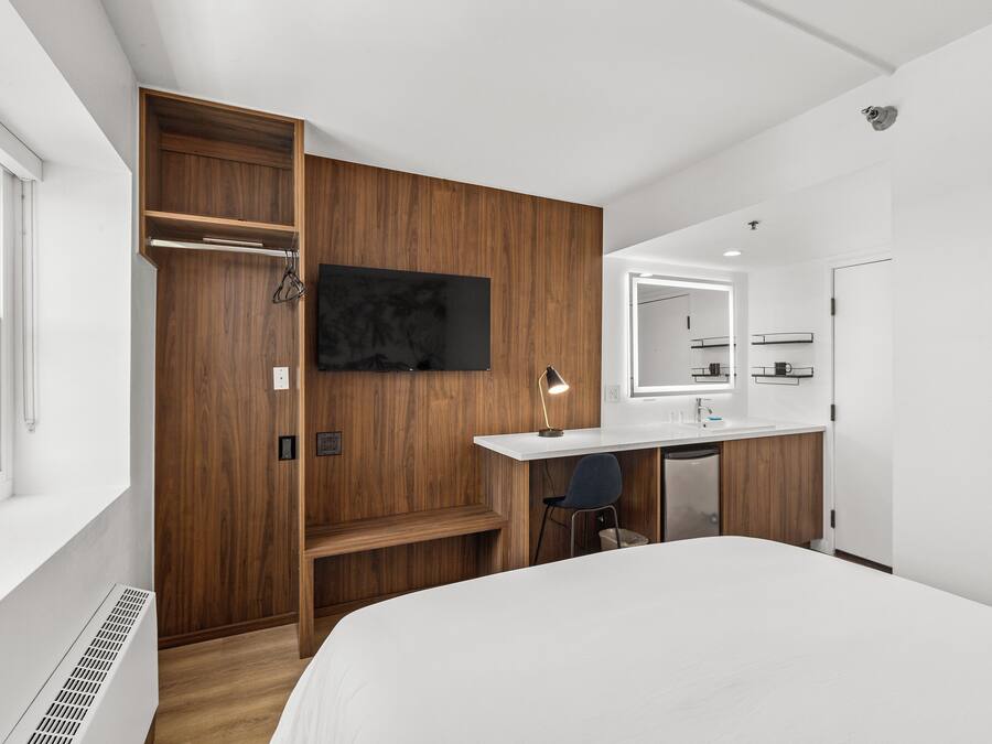 A modern hotel room with a wooden wardrobe, a bed, a desk with a chair, a TV, and a bathroom with a mirror and sink.