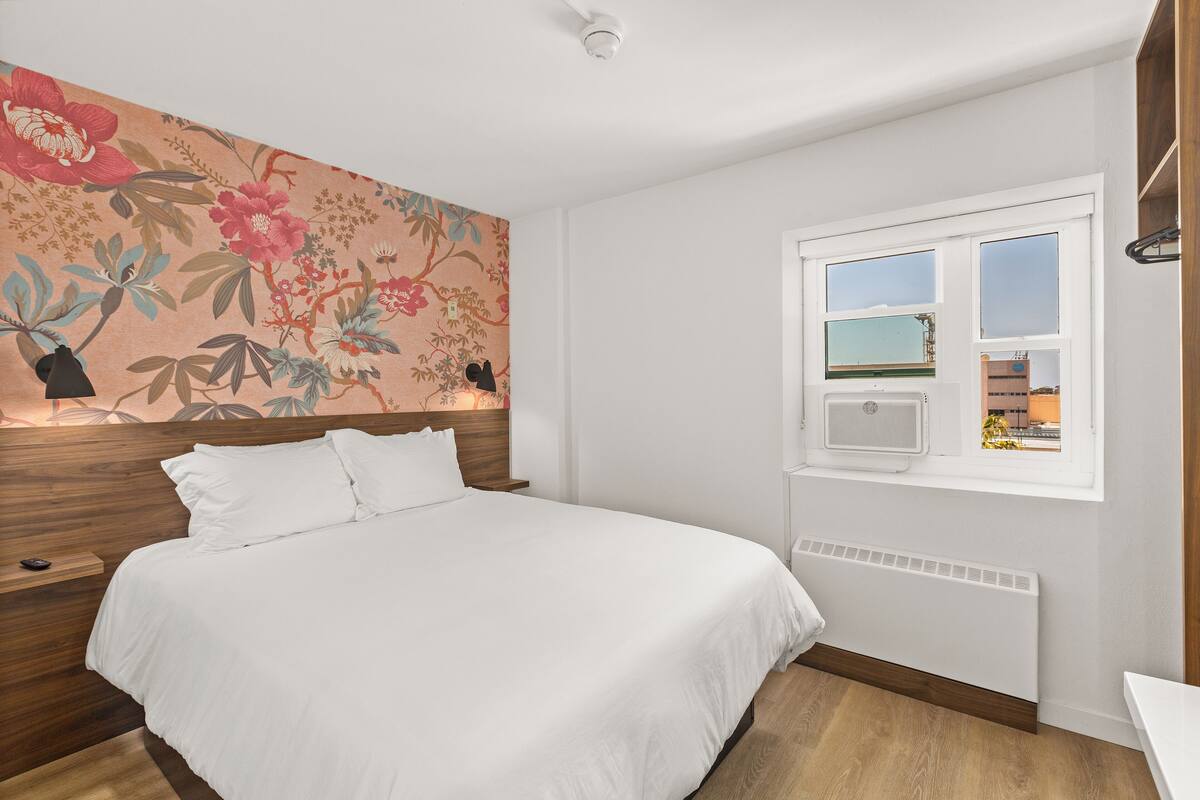 Ein Schlafzimmer mit einem großen Bett, einem Holzkopfbrett, einem blumigen Wandbild, einem Fenster mit Aussicht und einem weißen Nachttisch.