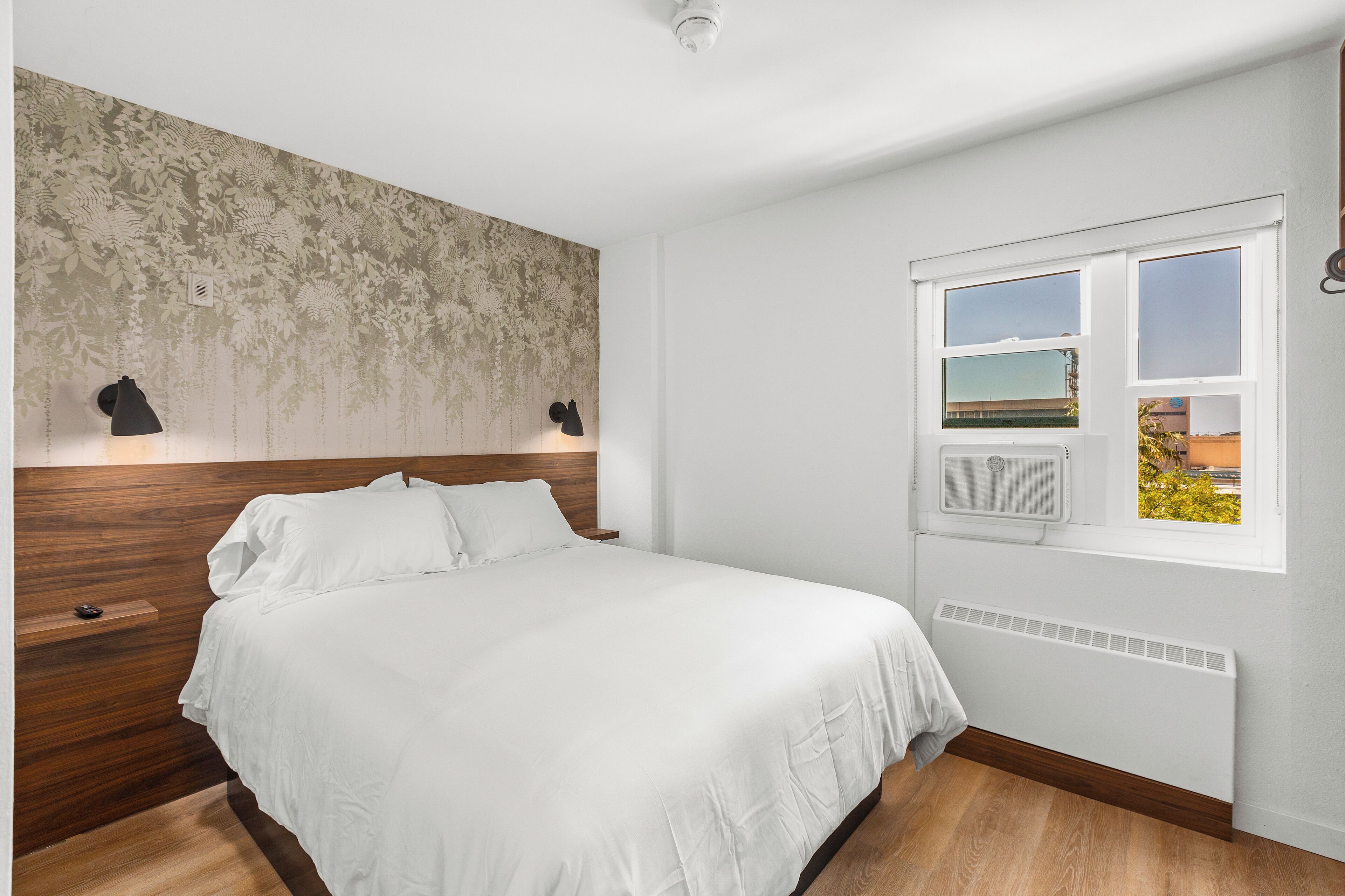 Un dormitorio con cabecera de madera, ropa de cama blanca, una lámpara de pared y una ventana con vistas.