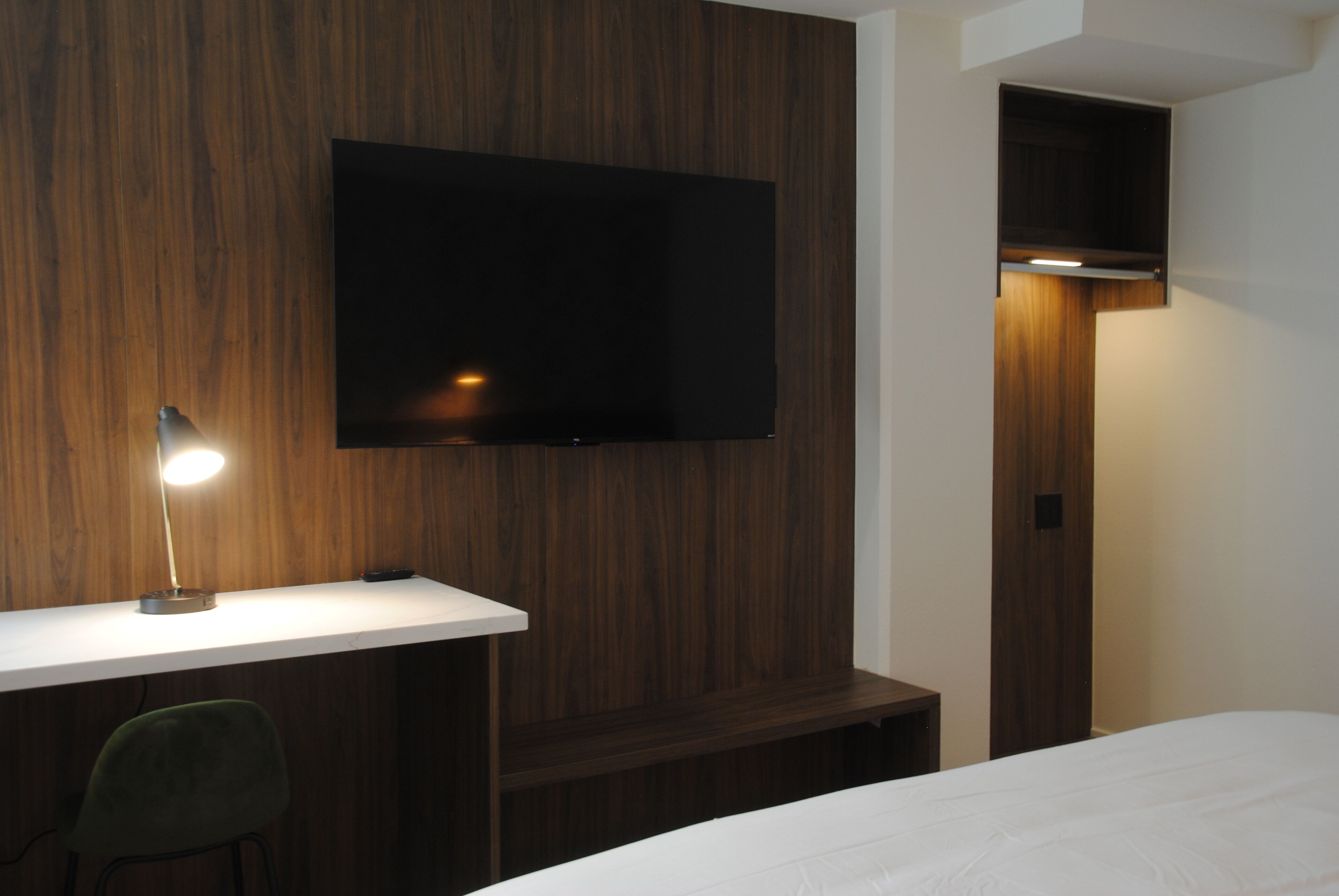 Habitación de hotel con pared de madera, un televisor de pantalla plana sobre un escritorio, una silla verde y una cama con ropa blanca.
