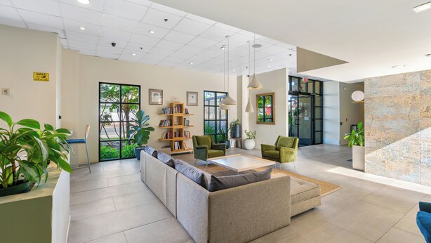 A spacious lobby with a sofa, chairs, a bookshelf, and a large window with a view of greenery. - Uptown Inn by Vantaggio - Limited Service Apart-Hotel (San Diego)