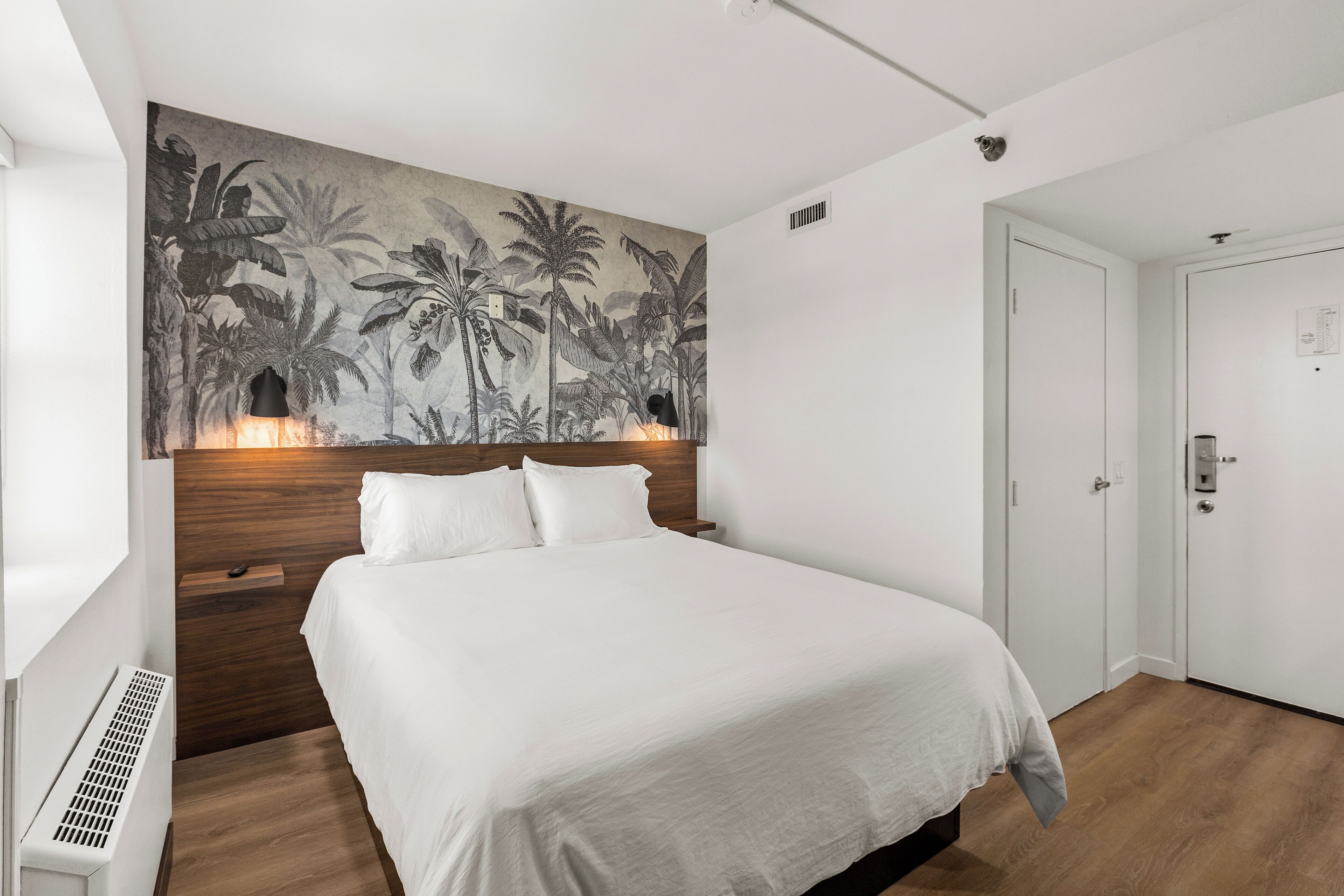 Une chambre d’hôtel avec un grand lit, une œuvre d’art représentant une végétation tropicale accrochée au mur, une porte blanche et un radiateur blanc.