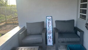 A covered outdoor seating area with two grey sofas, a wooden table, and a welcome sign.