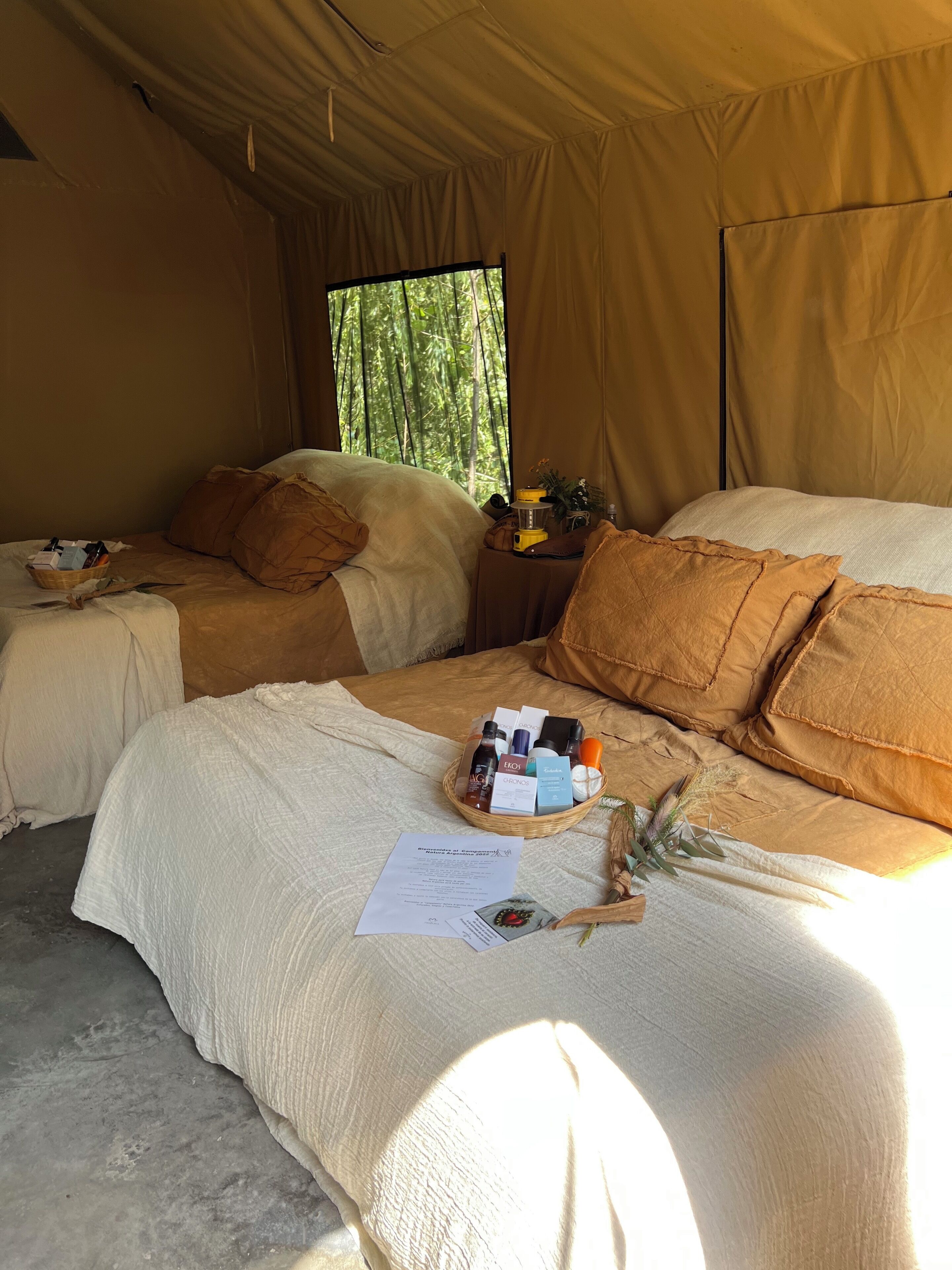 A tented accommodation with two beds, a small table with toiletries, and a window with a view of trees.