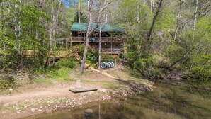 A cabin with a green roof and a deck, surrounded by trees and a small stream.