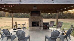 A covered outdoor seating area with a stone fireplace, surrounded by chairs and a fire pit.