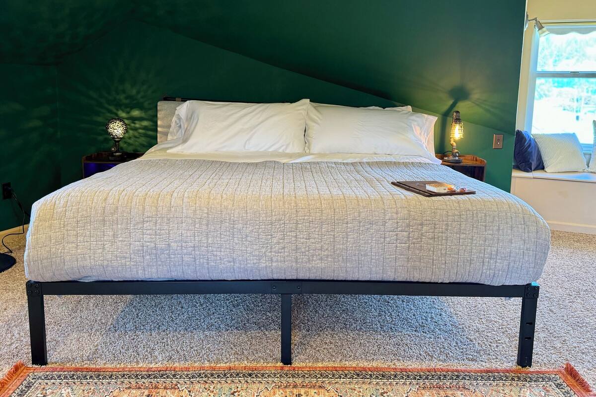 A bed with a quilted white cover, a bedside lamp, and a small table with a book and a tray.