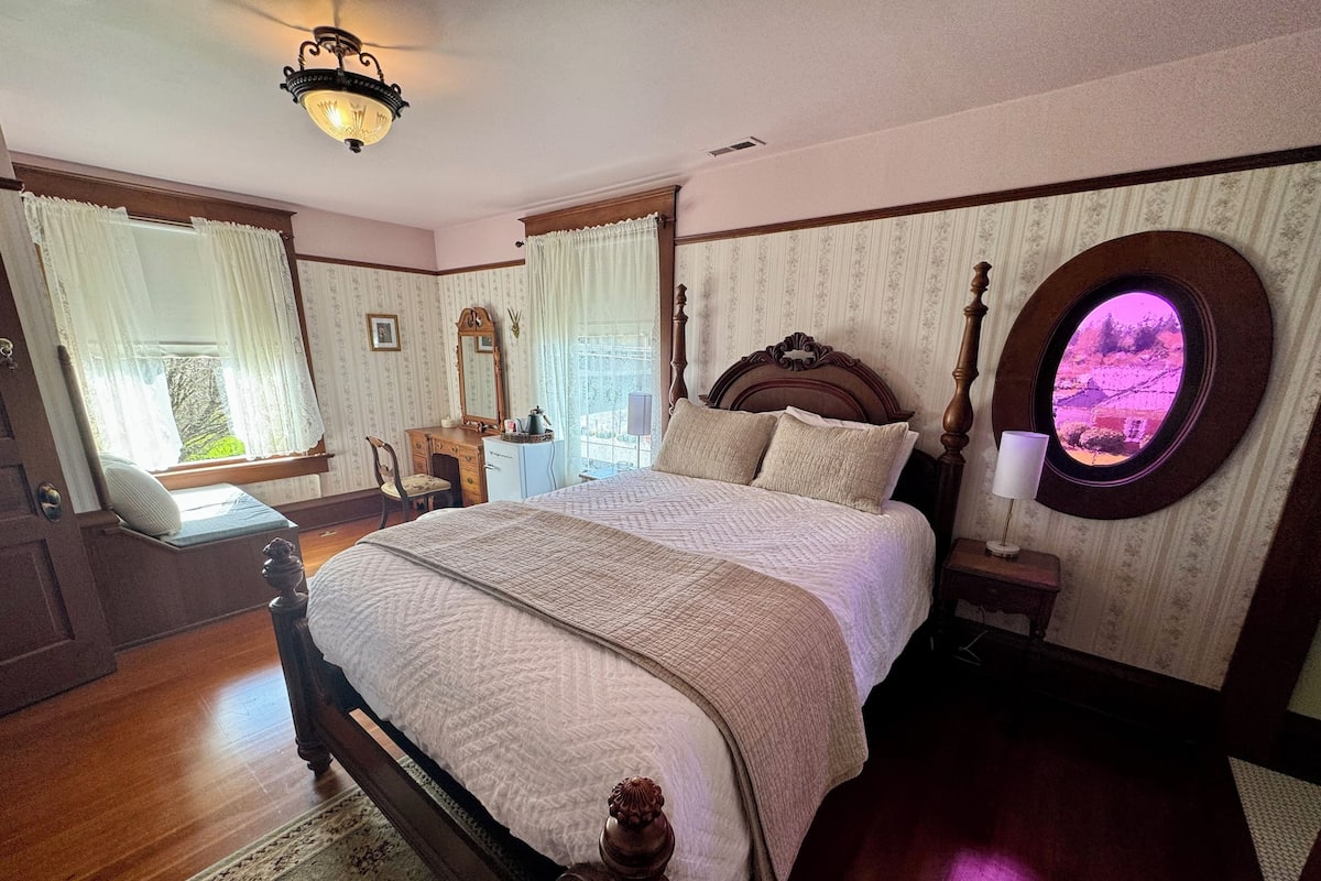 A traditional bedroom with a large bed, wooden headboard, a round mirror, and a view of a garden through a window.