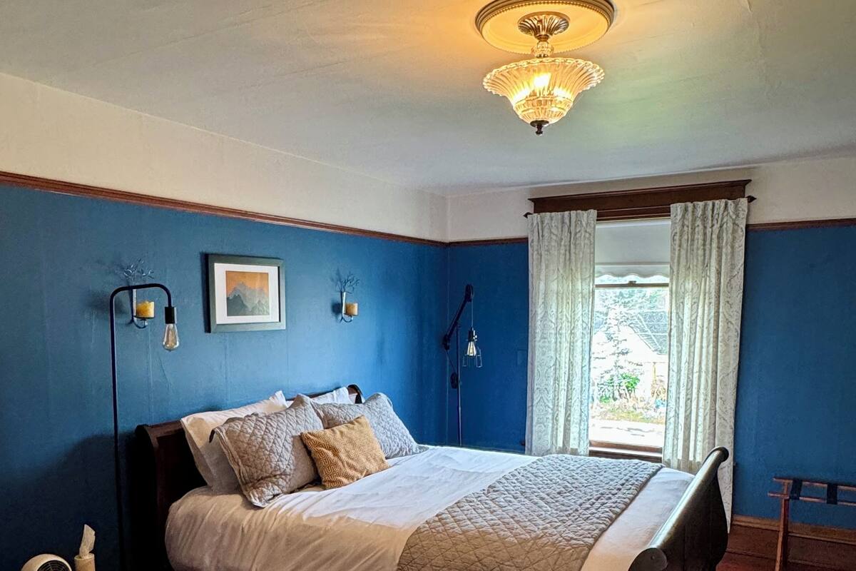 A bedroom with a bed, a nightstand, a lamp, a window with curtains, and a view of greenery outside.