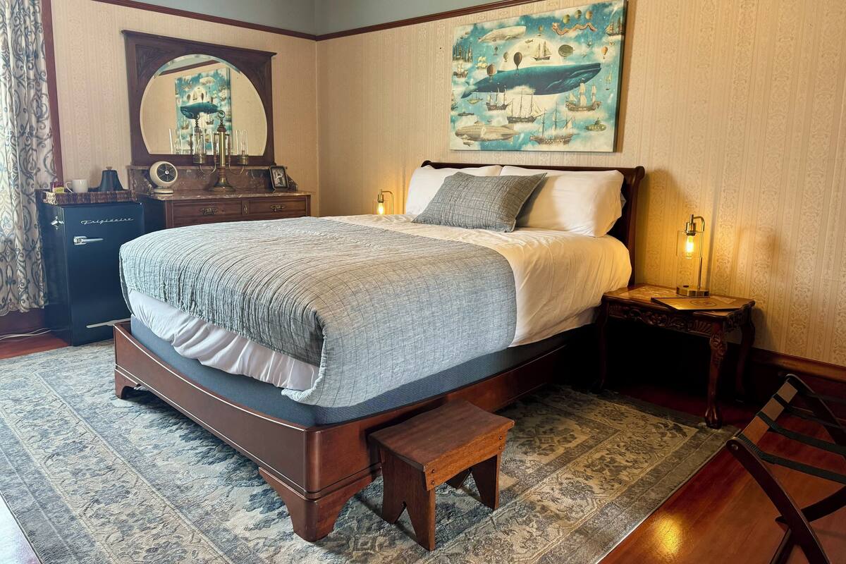 A bedroom with a large bed, a dresser, a mirror, a painting, and a bedside table with a lamp.
