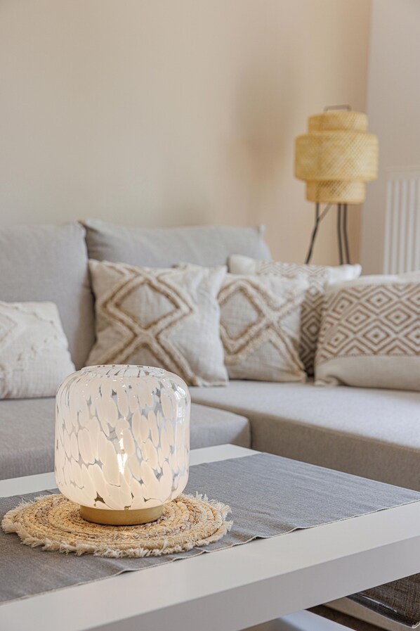 Una sala moderna con una lámpara de vidrio decorativa sobre un posavasos tejido, un sofá gris con cojines estampados y una lámpara amarilla al fondo.