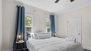 A bedroom with a large bed, white bedding, a small table with a lamp, and blue curtains. - Adorable Bungalow, Minutes from Downtown Durham (Durham)