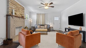 A living room with a brick fireplace, two orange armchairs, a grey sofa, a flat-screen TV, and a ceiling fan. - Adorable Bungalow, Minutes from Downtown Durham (Durham)