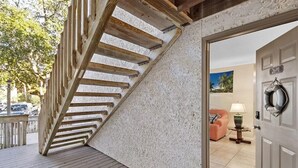 Un escalier en bois mène à une pièce dont le mur est recouvert d’un revêtement texturé blanc, où l’on peut voir une pancarte d’accueil et une porte portant une plaque avec un numéro.