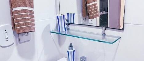 Un baño con un lavamanos blanco, un estante de vidrio y dos toallas colgadas en argollas.