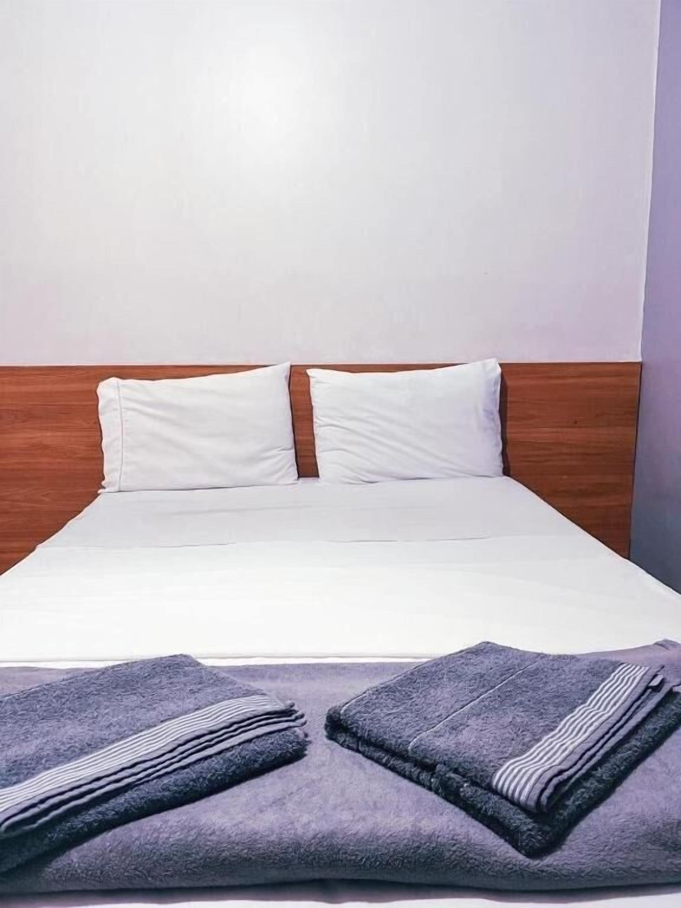 Cama bem arrumada com lençóis brancos e duas almofadas, cabeceira de madeira e duas toalhas dobradas sobre a cama.