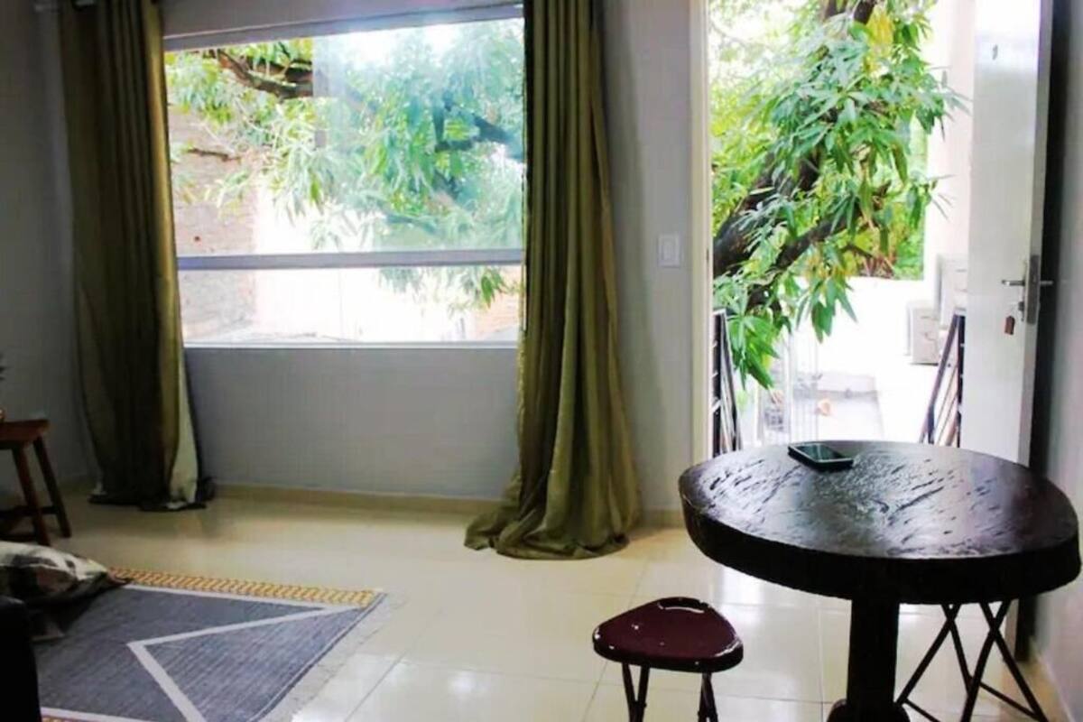 A room with a large window, a round table with a chair, a small stool, and a plant.