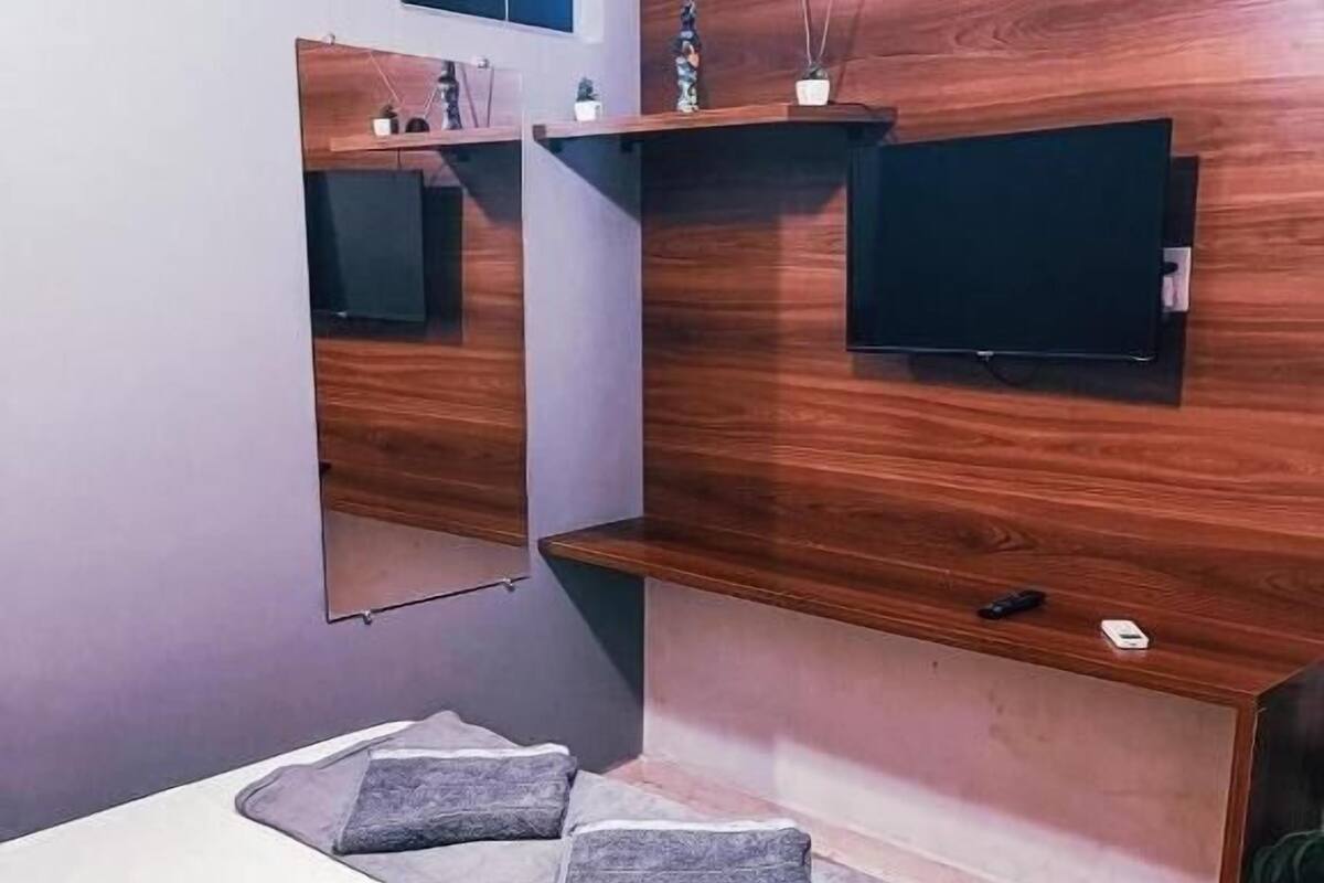 A modern bedroom with a bed, a TV mounted on a wooden wall, and a small shelf with a remote control.