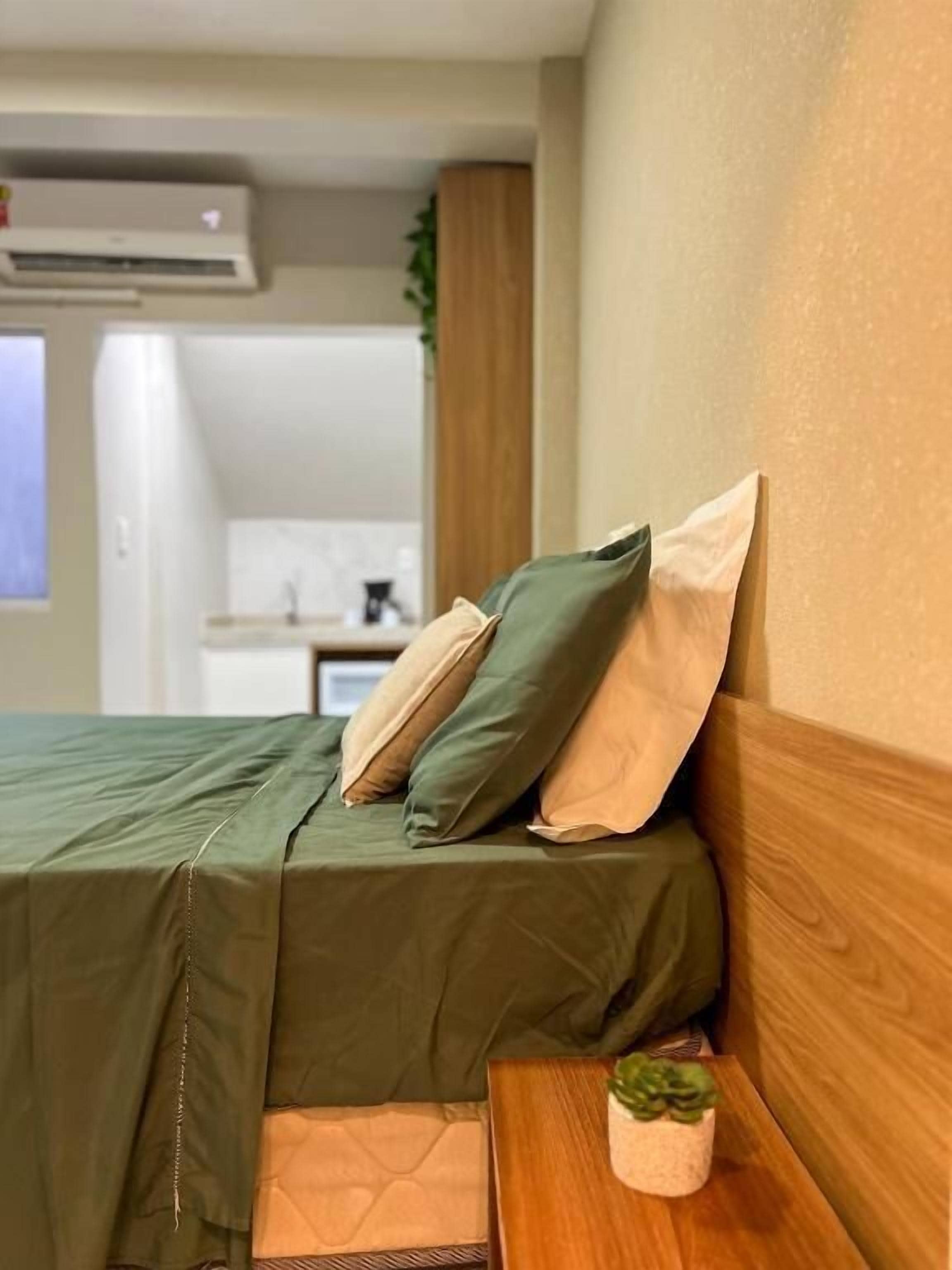 A neatly made bed with a green bedspread and two pillows, a wooden headboard, and a small bedside table with a potted plant.