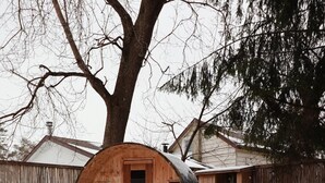 Een besneeuwde buitenruimte met Adirondack-stoelen en een vuurkuil, omringd door een houten vatstructuur en een houten hek.
