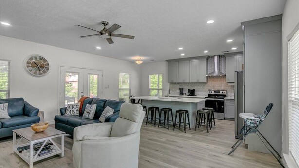 A modern living room with a sofa, a coffee table, and a white armchair. There is a kitchen area with a stove, sink, and bar stools. - Waterfront house on Sam Rayburn (Brookeland)
