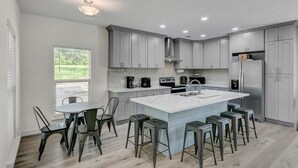 A modern kitchen with grey cabinets, a large island with bar stools, a dining table with chairs, and a window with a view of greenery. - Waterfront house on Sam Rayburn (Brookeland)