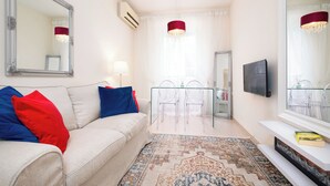 A modern living room with a sofa, red and blue cushions, a glass coffee table, a patterned rug, a large mirror, a TV, and a chandelier. - Guilford Elegance | Corfu Old  town Apartment | fast WiFi (Corfu)