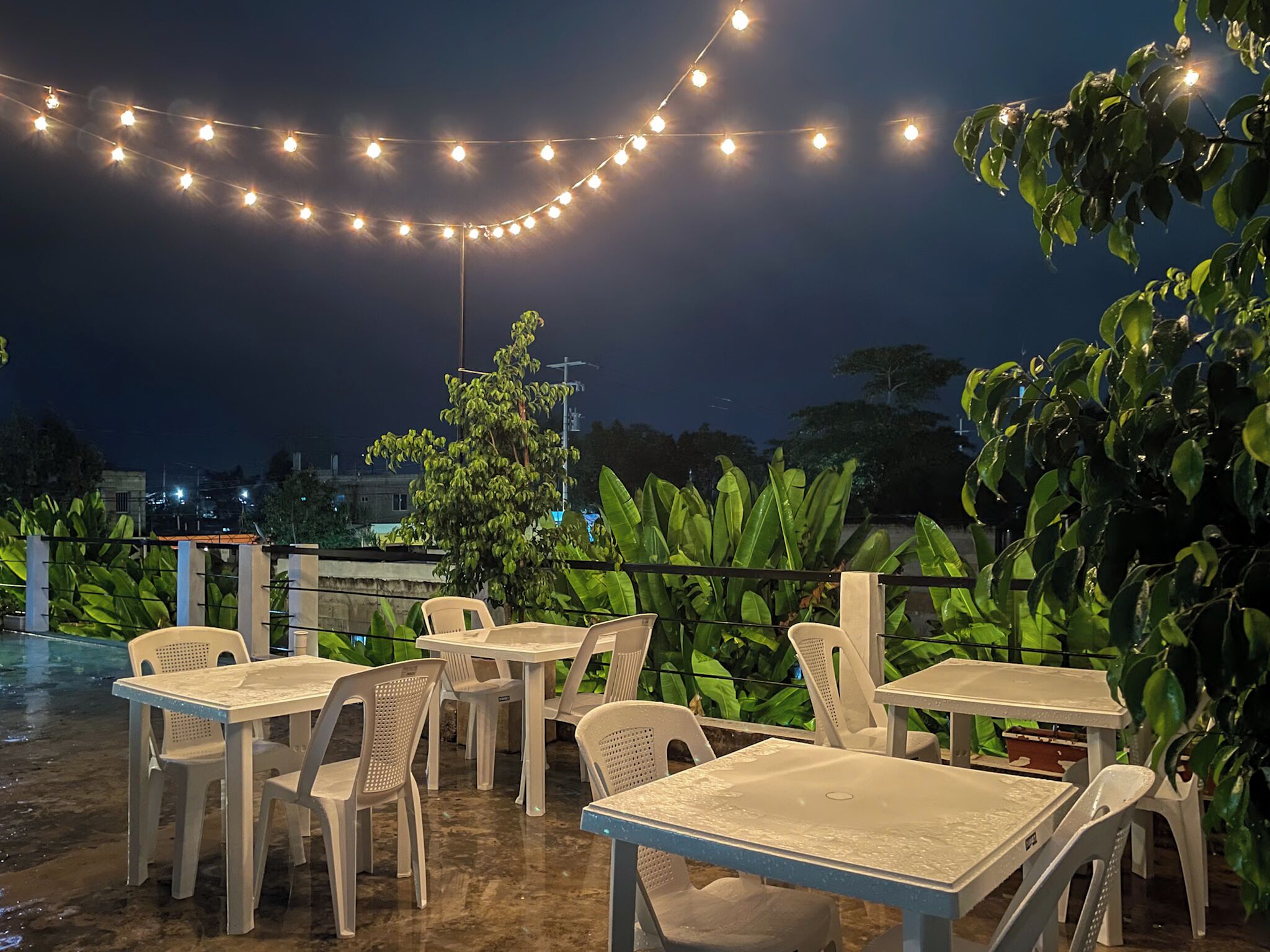 An outdoor seating area with tables and chairs, string lights overhead, and lush greenery in the background.