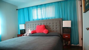 A bedroom with a large bed, a tufted headboard, and a bright blue window with blinds. - The Big Apple City View with Balcony (Dayton)