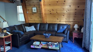 Una sala de estar acogedora con techo de madera, un sofá de cuero negro, una mesa de centro con juegos de mesa y una escalera que conduce a un nivel superior.