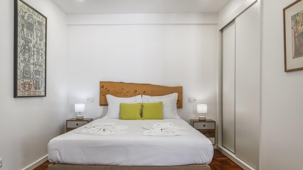 Un lit bien fait, avec du linge de lit blanc et deux oreillers, une tête de lit en bois, des tables de chevet avec des lampes et des œuvres d’art accrochées aux murs.