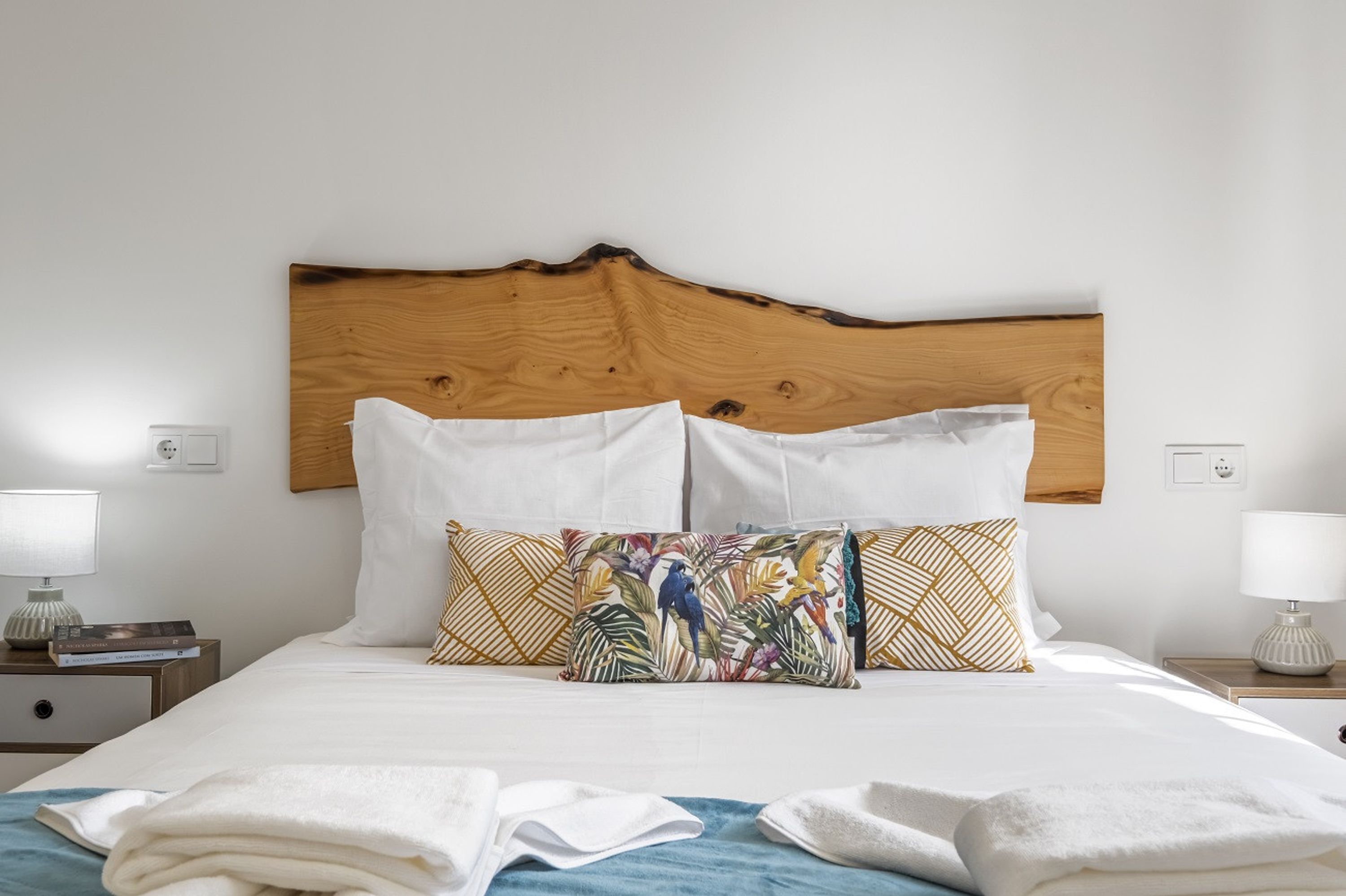 A neatly made bed with white linens and two decorative pillows, one with a tropical print and the other with a geometric pattern.
