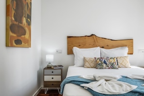 Een netjes opgemaakt bed met witte lakens en een blauwe dekbedovertrek, een houten hoofdbord, en een nachtkastje met een lamp en een boek.