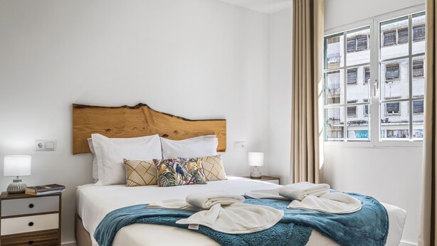 Ein ordentlich bezogenes Bett mit Holz-Kopfstütze, weißer Bettwäsche und einer blauen Tagesdecke. Zwei Nachttische mit Lampen und einer kleinen Schublade sind vorhanden.