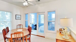 A spacious room with a round wooden dining table, six chairs, a ceiling fan, and a lamp on a wooden side table. - Mercy Me by Tybee Vacation Rentals (Tybee Island)