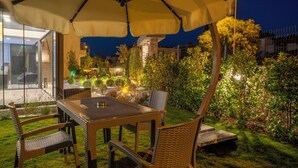 Un espace extérieur aménagé pour les repas, avec une table et des chaises placées sous un grand parasol, entouré d’une végétation bien entretenue et agrémenté, au fond, d’une structure en pierre.