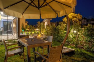 Un área de comedor al aire libre con una mesa y sillas bajo un paraguas grande, rodeada de vegetación cuidada y una estructura de piedra al fondo.