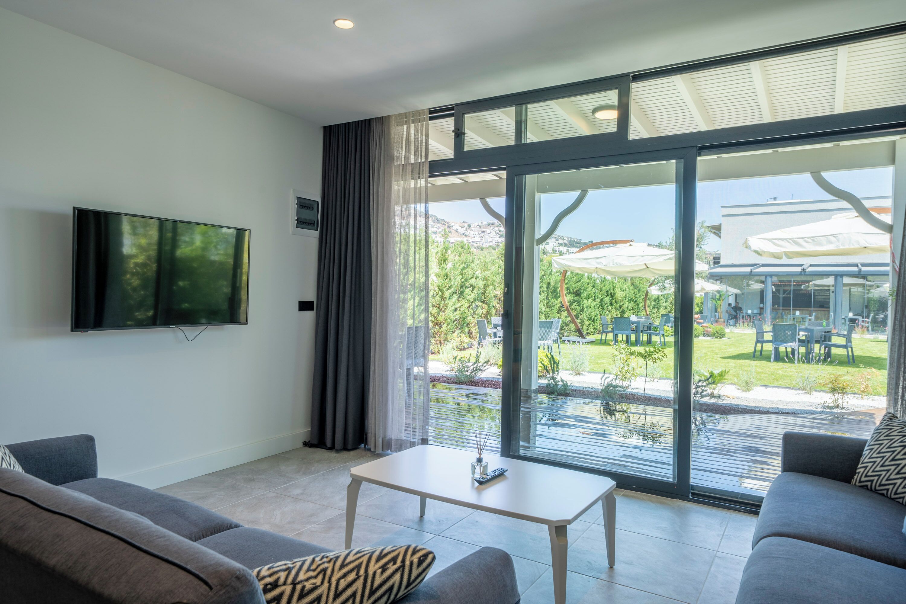 A modern living room with a flat-screen TV, two sofas, a coffee table, and a large sliding glass door leading to an outdoor area with umbrellas and seating.