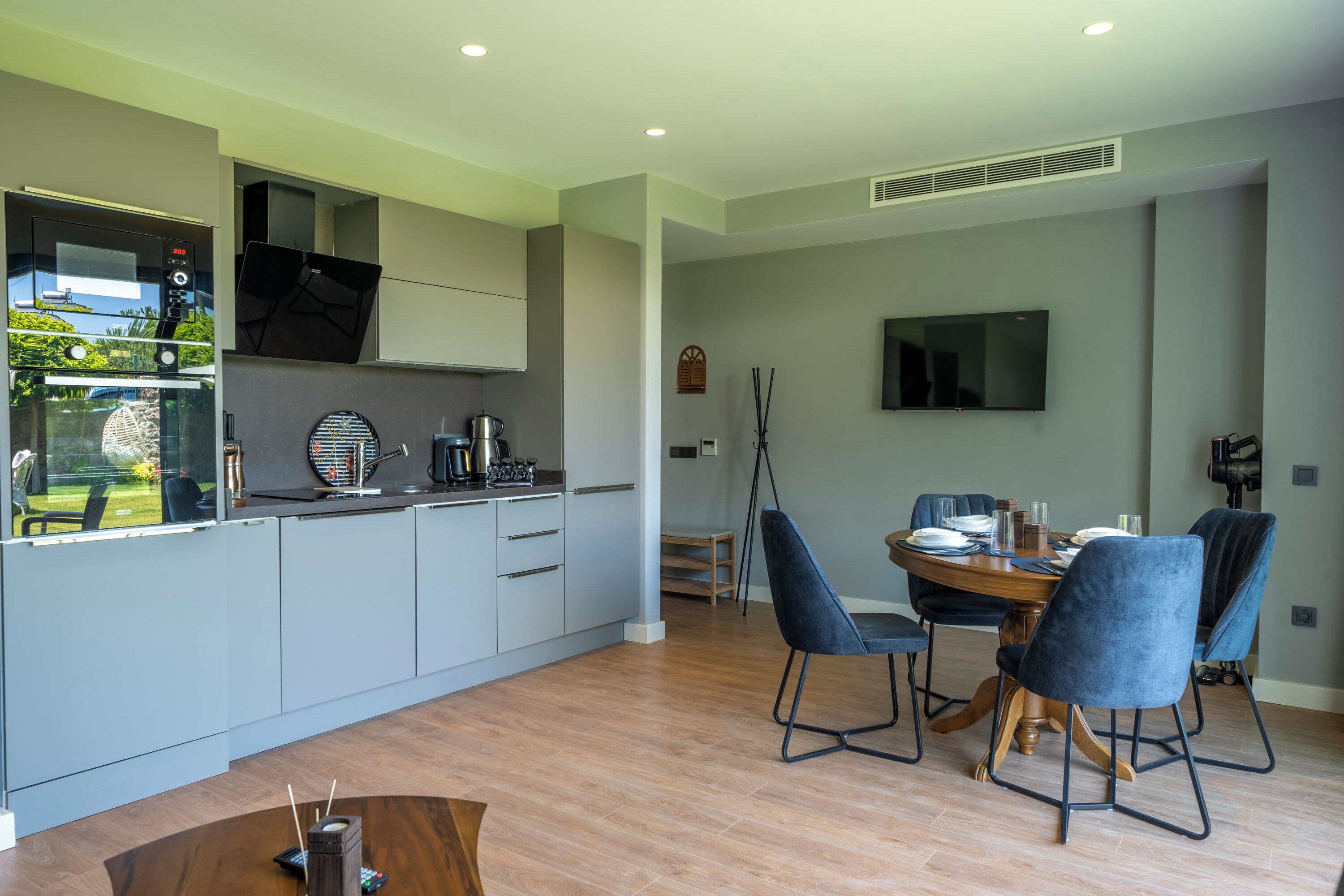 A modern kitchen with grey cabinets, a built-in oven, and a dining area with a round table and blue chairs.