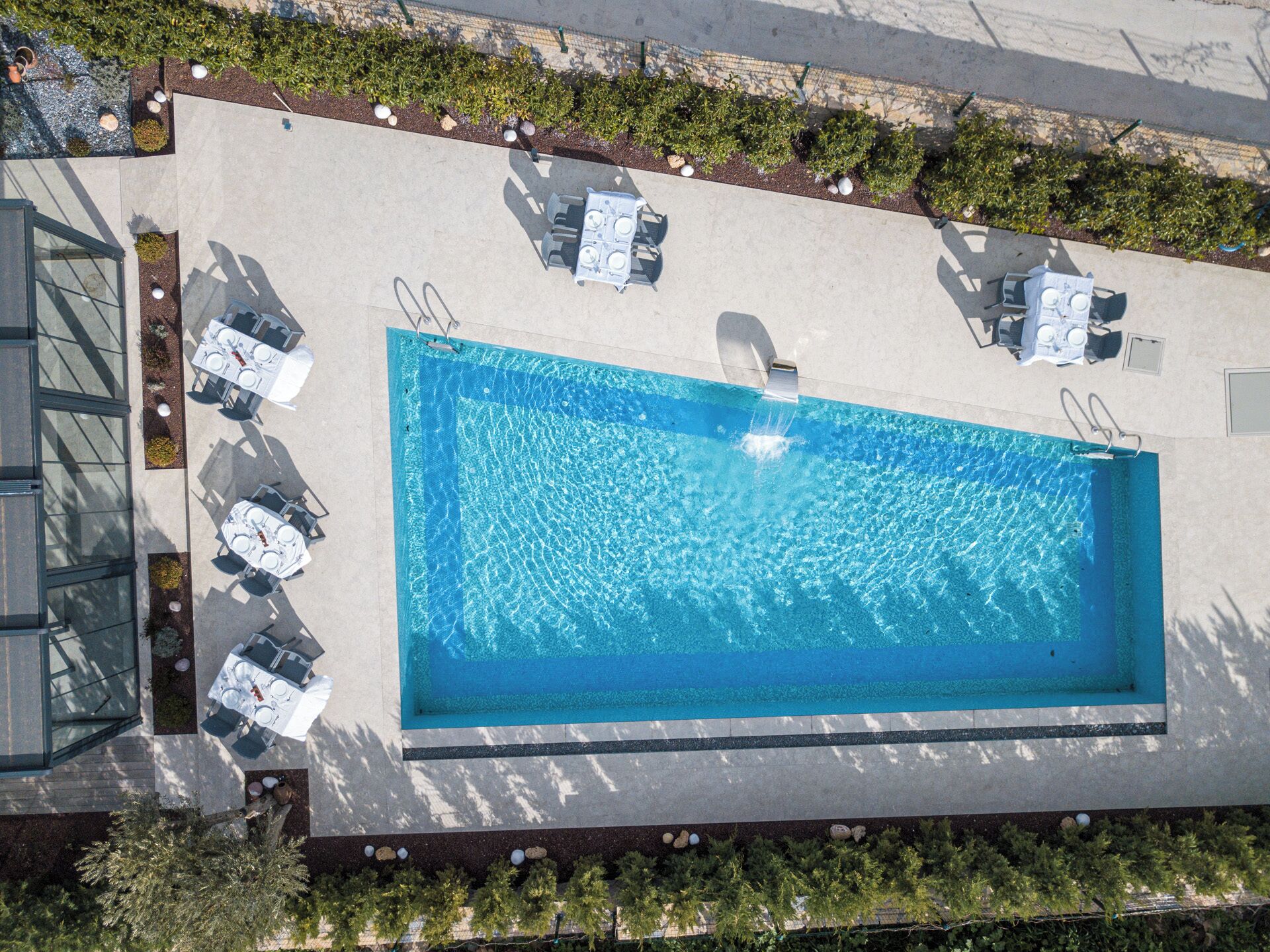 Vista aèria d'una piscina rectangular amb superfície de blau clar, envoltada per un solàrium de formigó. Hi ha quatre gandules blanques i una taula petita amb una cadira al solàrium.