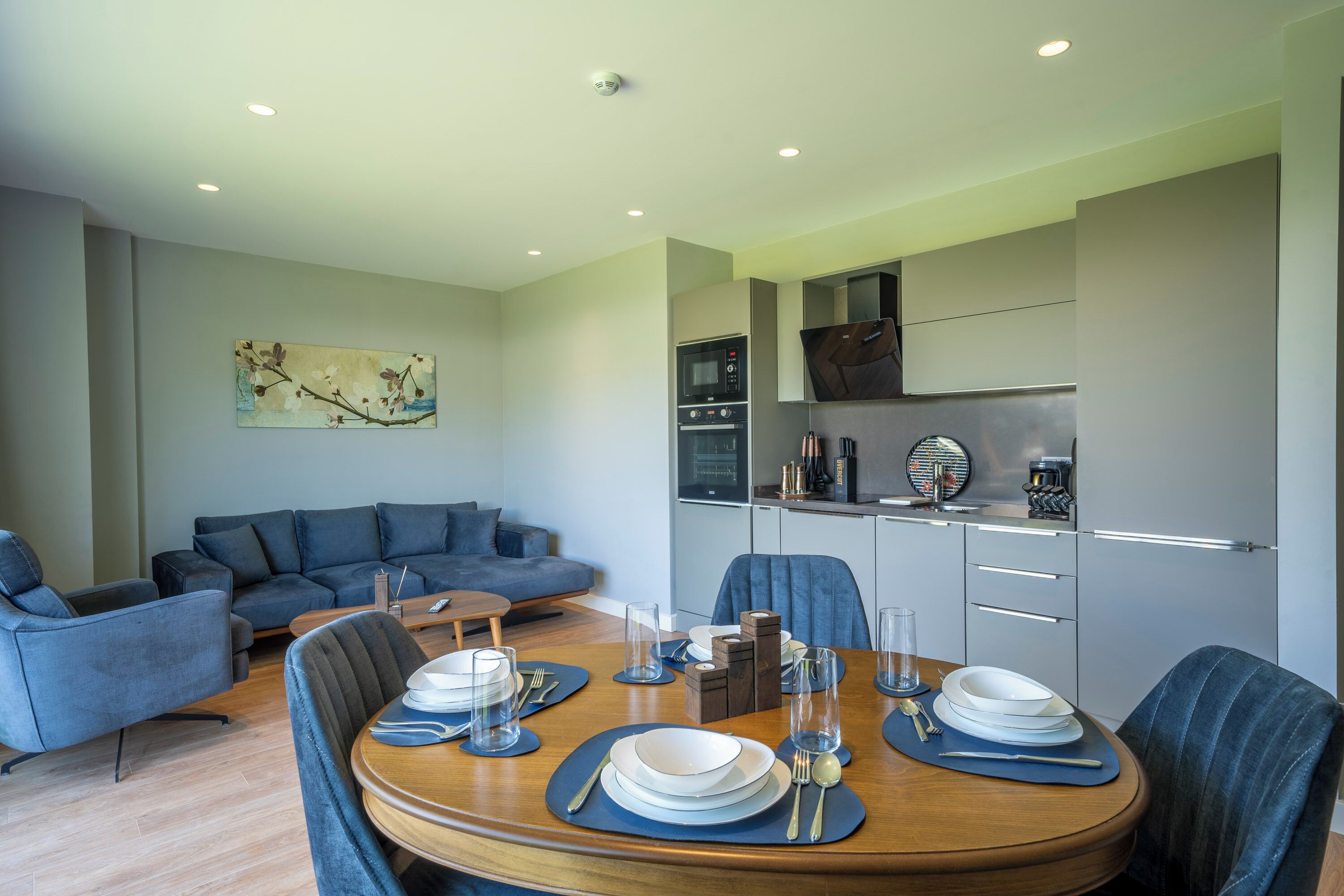 A modern living room with a sofa, a dining table set for four, and a kitchen area with appliances.