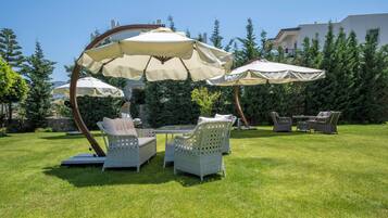 Eine Terrasse mit Korbmöbeln und großen Sonnenschirmen, umgeben von viel Grün und einem Gebäude im Hintergrund.
