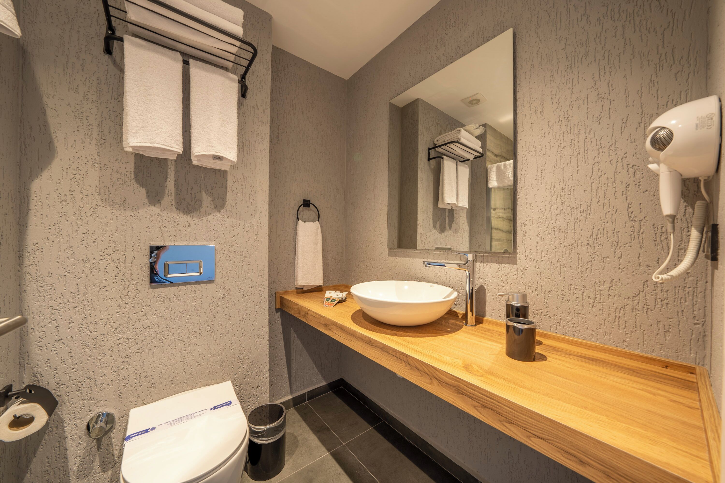 A modern bathroom with a wooden vanity, a large mirror, a white sink, and a hairdryer mounted on the wall.