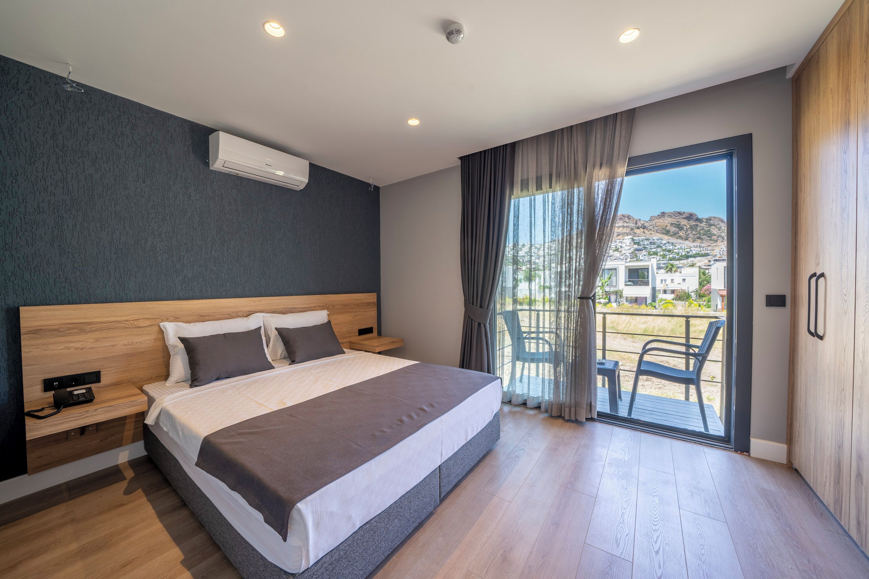 Quarto de hotel moderno com uma cama grande, cabeceira de madeira, mesa de cabeceira e vista para a varanda externa.