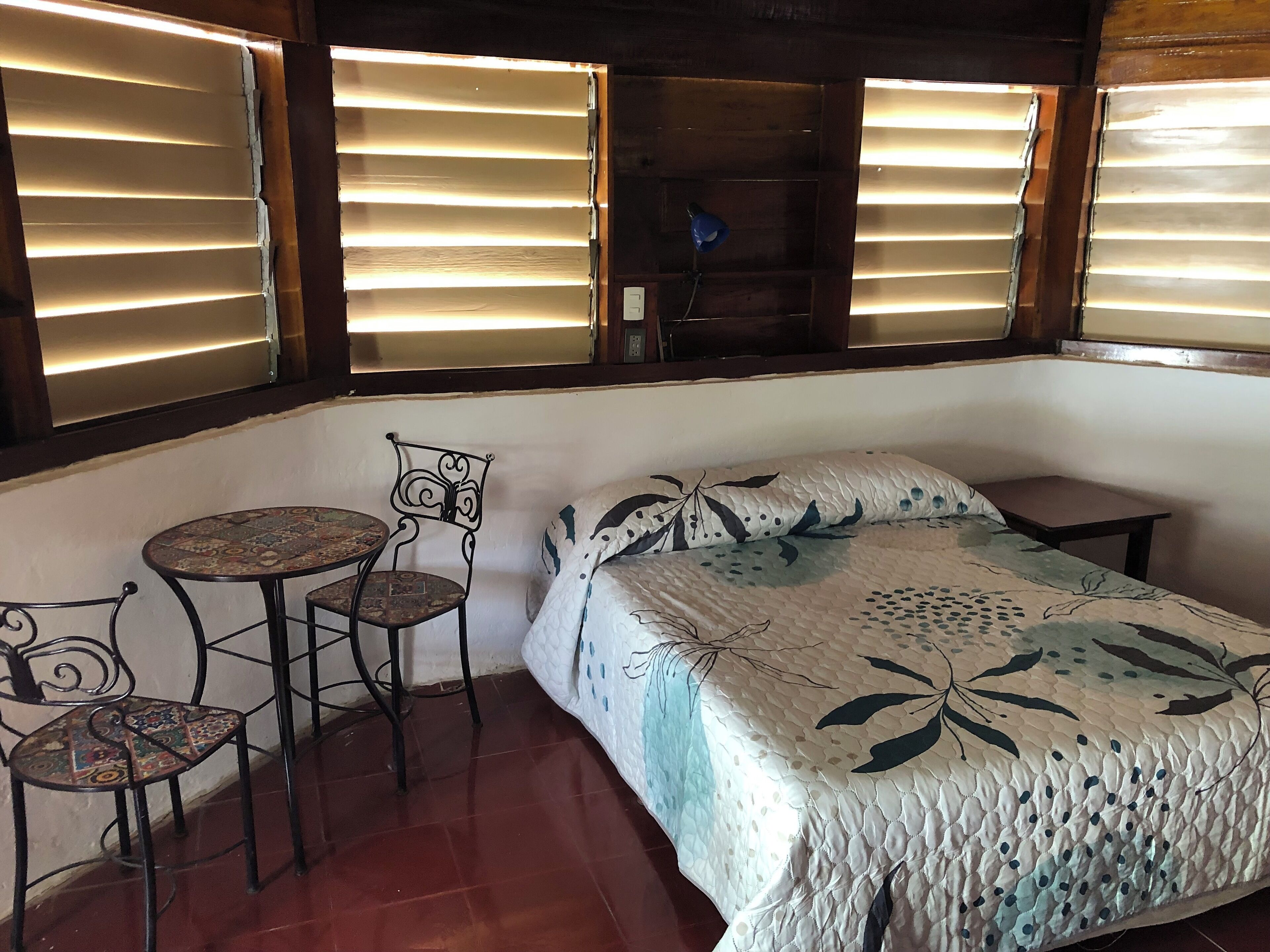 Ein Schlafzimmer mit einem Bett, einem kleinen Tisch, zwei Stühlen und einem Fenster mit Jalousien.