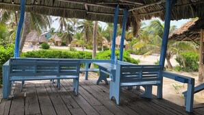 Un pontile in legno con panchine sotto un tetto di paglia, circondato da palme e una spiaggia sullo sfondo.